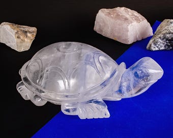 May include: A clear quartz crystal turtle sculpture with detailed carving, displayed on a blue and black surface. Several raw quartz stones in shades of white, gray, and tan are arranged around the turtle.