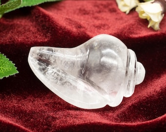 Puede incluir: Un cristal de cuarzo transparente tallado en forma de concha. El cristal es liso y pulido, y está descansando sobre una superficie de terciopelo rojo.