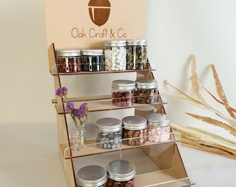 May include: Wooden display stand with multiple shelves showcasing small glass jars filled with colourful wax beads. The stand has a sign with the text "Oak Craft & Co" and an acorn logo.