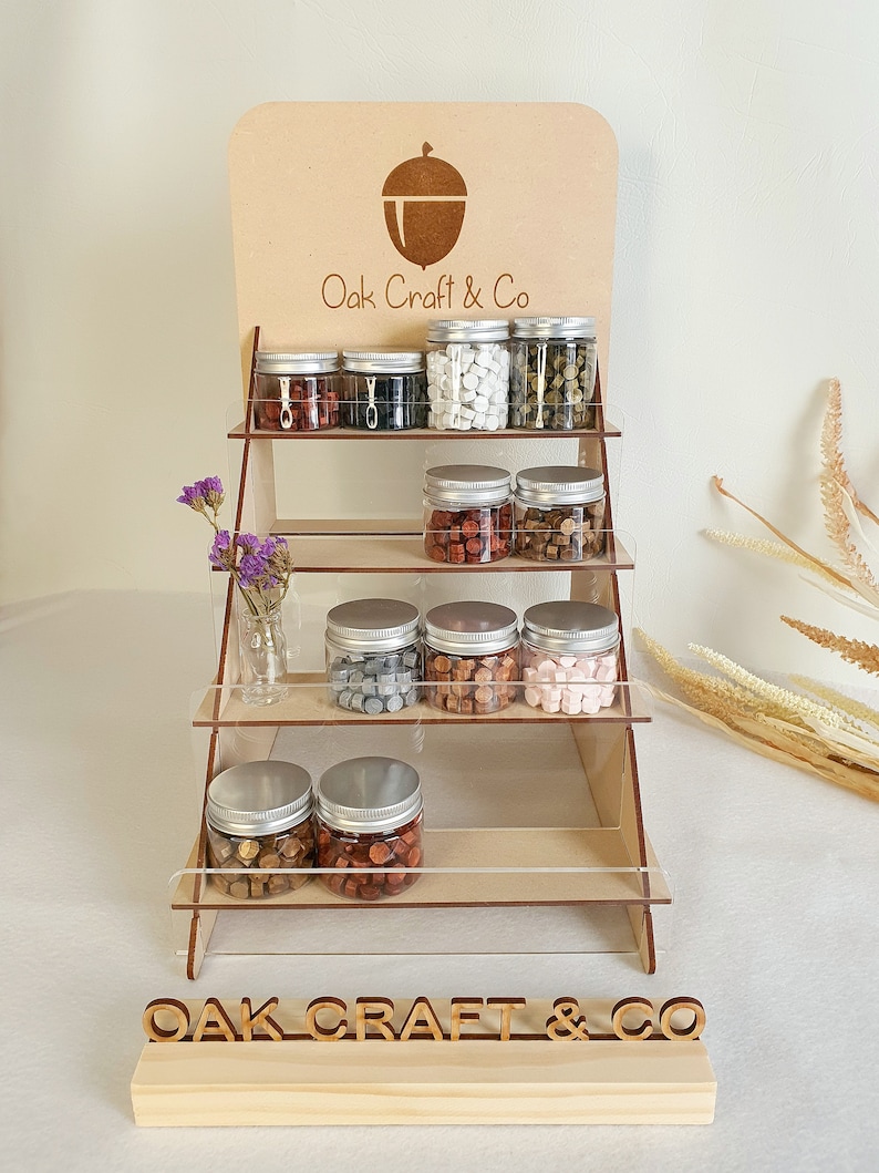May include: A wooden display stand with four shelves, showcasing various colours of sealing wax in glass jars. The stand has a sign at the top that reads "Oak Craft & Co." and a smaller sign at the bottom that reads "OAK CRAFT & CO."