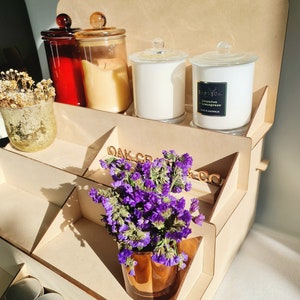 May include: A wooden tiered display stand with a variety of candles and a small bouquet of purple flowers in a brown glass vase. The stand has the words "Oak Creek Co." printed on the side.