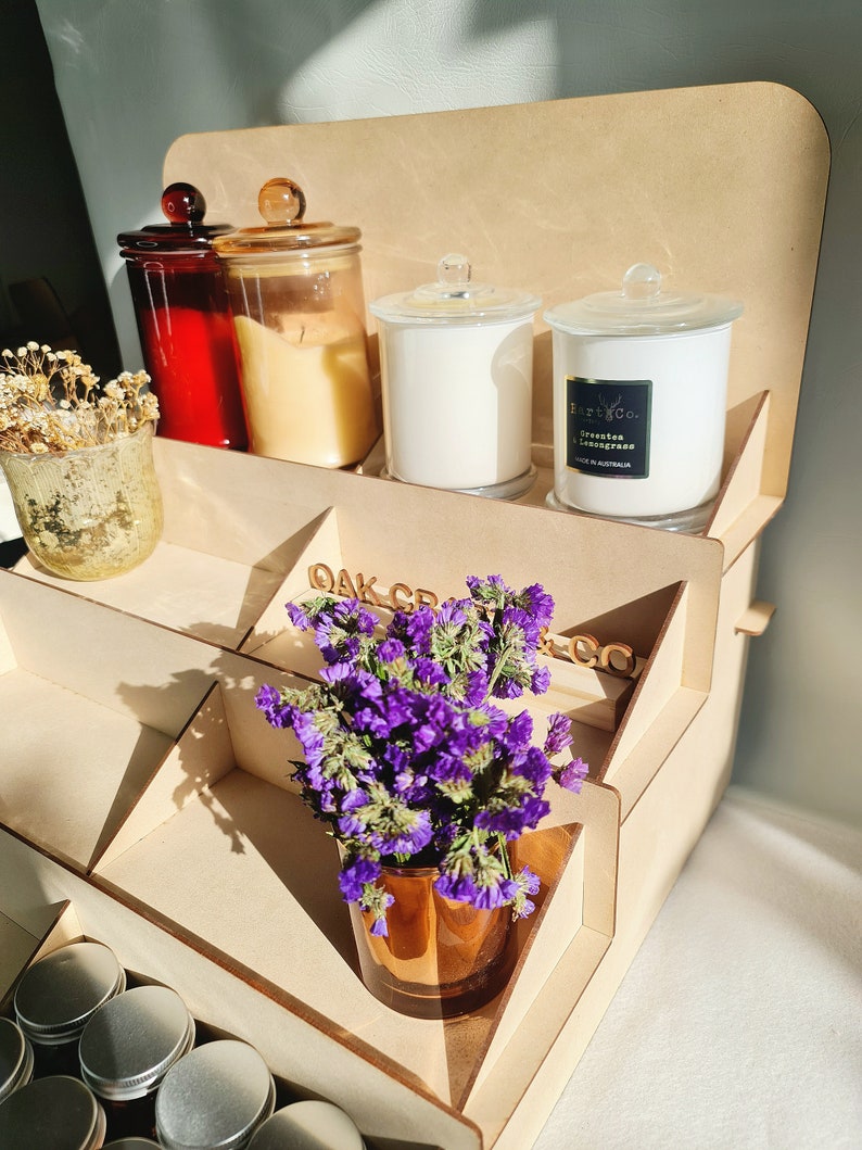 May include: A wooden tiered display stand with a variety of candles and a small bouquet of purple flowers in a brown glass vase. The stand has the words "Oak Creek Co." printed on the side.