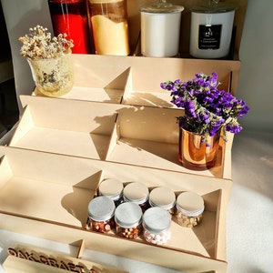 May include: A wooden tiered display stand with a variety of items, including candles, jars, and small containers. The stand is made of natural wood and has a rustic look. The top tier features a white candle with a gold lid, a clear glass jar with a gold lid, and a red glass jar with a gold lid. The middle tier features a vase with dried flowers and a clear glass jar with a gold lid. The bottom tier features a row of small containers with silver lids, filled with colorful wax melts. The stand is labeled "Oak Craft & Co."