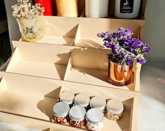 May include: A wooden tiered display stand featuring a selection of items, including candles, jars, and small containers. The stand is crafted from natural wood and has a rustic appearance. The top tier showcases a white candle with a gold lid, a clear glass jar with a gold lid, and a red glass jar with a gold lid. The middle tier displays a vase with dried flowers and a clear glass jar with a gold lid. The bottom tier features a row of small containers with silver lids, filled with colourful wax melts. The stand is labelled "Oak Craft & Co."