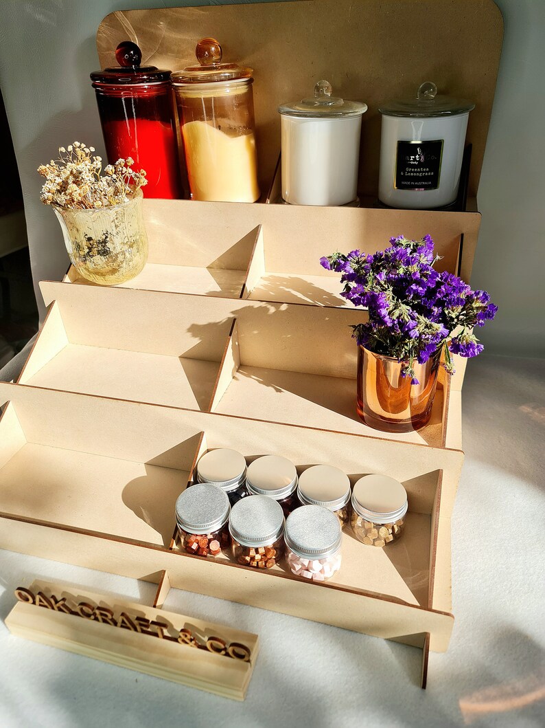 May include: A wooden tiered display stand with a variety of items, including candles, jars, and small containers. The stand is made of natural wood and has a rustic look. The top tier features a white candle with a gold lid, a clear glass jar with a gold lid, and a red glass jar with a gold lid. The middle tier features a vase with dried flowers and a clear glass jar with a gold lid. The bottom tier features a row of small containers with silver lids, filled with colorful wax melts. The stand is labeled "Oak Craft & Co."