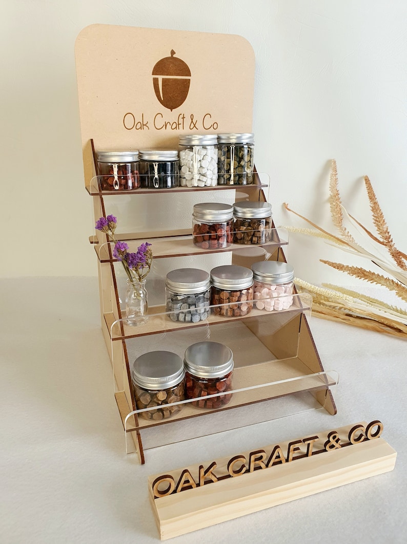 May include: Wooden display stand with multiple shelves showcasing small glass jars filled with colourful wax beads. The stand has a sign with the text "Oak Craft & Co" and an acorn logo.