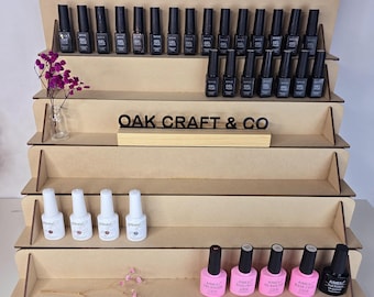 May include: A multi-tiered, light brown wooden display stand holding various nail polish bottles. The stand features the text "OAK CRAFT & CO." and is decorated with small floral arrangements. The nail polish bottles are in various colours.