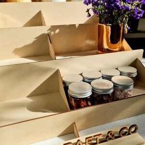 May include: A wooden tiered display stand with a sign that reads "OAK CRAFT & CO". The stand is filled with small glass jars with silver lids, filled with various items. A vase with purple flowers sits on the top shelf.