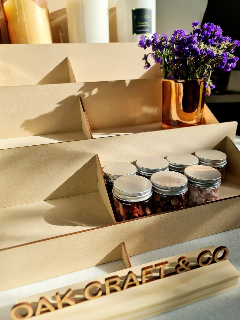 May include: A wooden tiered display stand with a sign that reads "OAK CRAFT & CO". The stand is filled with small glass jars with silver lids, filled with various items. A vase with purple flowers sits on the top shelf.