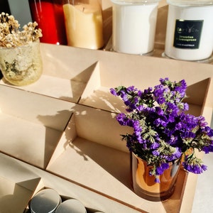 May include: A wooden shelf unit with multiple compartments, displaying a variety of items, including candles, jars, and a small bouquet of purple flowers in a copper vase. The candles are in glass jars with lids, and the jars are in various colors, including red, brown, and clear. The shelf unit is made of light wood and has a natural finish.