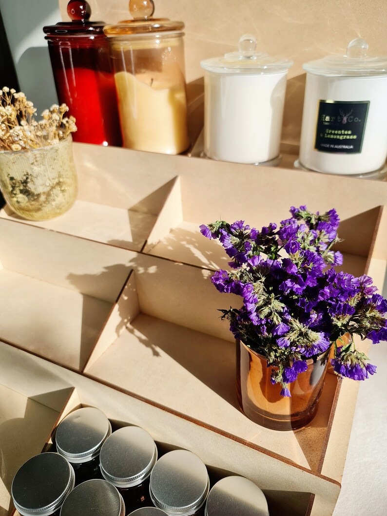 May include: A wooden shelf unit with multiple compartments, displaying a variety of items, including candles, jars, and a small bouquet of purple flowers in a copper vase. The candles are in glass jars with lids, and the jars are in various colors, including red, brown, and clear. The shelf unit is made of light wood and has a natural finish.