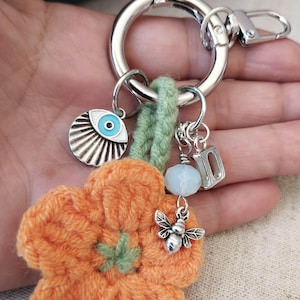 May include: A silver-toned metal keychain featuring a crocheted orange flower with a green center. Attached are a silver-toned evil eye charm with blue and white enamel, a small silver bee charm, a translucent white bead, and a silver letter D charm.