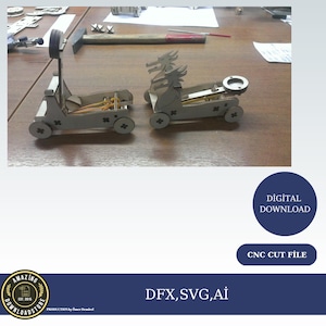 May include: Two wooden model catapults with rubber band launching mechanisms. One has a dragon head design. The models are on a wooden surface. The image also includes the text "DIGITAL DOWNLOAD" and "CNC CUT FILE".