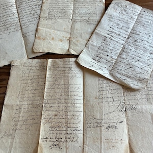 Könnte beinhalten: Vier antike, cremefarbene Pergamentdokumente mit handschriftlichem kursivem Text in dunkler Tinte. Die Papiere weisen sichtbare Knicke, Falten und altersbedingte Verfärbungen auf, wobei einige Abschnitte offizielle Stempel und Unterschriften zeigen.