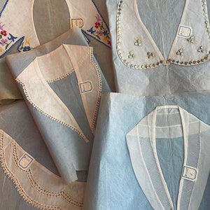 Può includere: Quattro colli bianchi con diversi ricami, tra cui pizzo, ricamo e paillettes, sono esposti su carta velina blu. I colli sono etichettati con etichette scritte a mano.