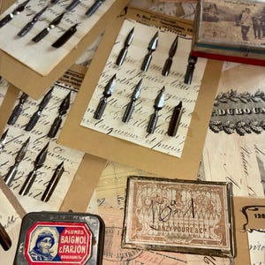 May include: A collection of antique dip pen nibs and ink boxes. The nibs are displayed on aged paper, some still on their original cards. Several small metal ink boxes are also present, featuring faded labels and illustrations.