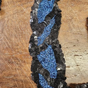 May include: Black and blue sequined trim with a wavy pattern. The trim is made of small, round sequins sewn together in a repeating pattern.