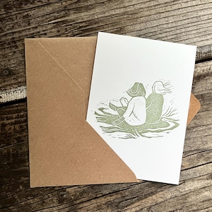 May include: A green and white greeting card with an illustration of two people sitting on a grassy surface. The people are facing away from the viewer and are drawn in a simple, blocky style.