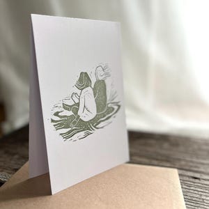 May include: A white card with a green ink illustration of two people sitting on a rock. The people are facing away from each other. The card is folded in half and sitting on a brown envelope.