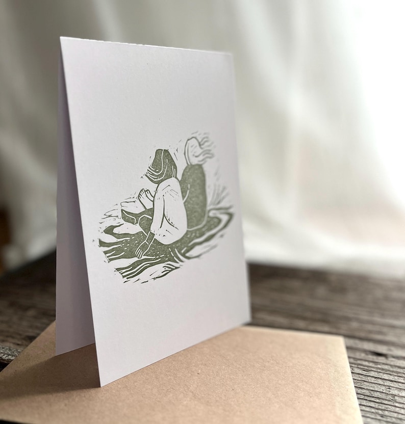 May include: A white card with a green ink illustration of two people sitting on a rock. The people are facing away from each other. The card is folded in half and sitting on a brown envelope.