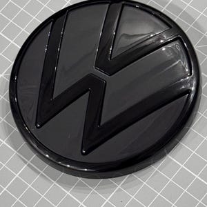 Op de afbeelding: Een glanzend zwart auto-embleem met het Volkswagen-logo. Het embleem is cirkelvormig met een verhoogd, gestileerd "VW"-ontwerp in het midden. Het embleem staat tegen een grijze rasterachtergrond.