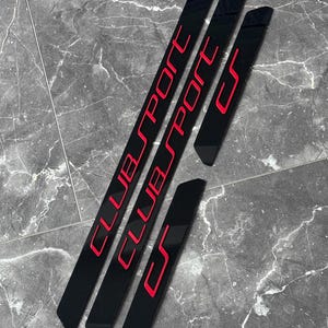 Könnte beinhalten: Schwarze Einstiegsleisten für Autotüren mit roter Schrift. Der Text lautet "CLUB SPORT" und "CS". Die Protektoren sind rechteckig und an den Enden abgewinkelt. Die Artikel sind auf einer grauen, strukturierten Oberfläche angeordnet.