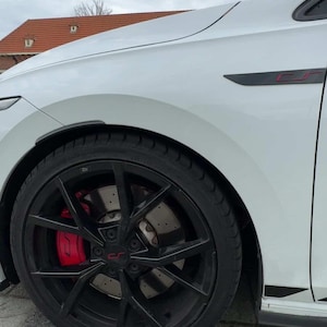Könnte beinhalten: Ein weißes Auto mit einem schwarzen Rad und roten Bremssätteln. Das Rad hat ein Mehrspeichen-Design. Das Auto hat einen schwarzen Akzent an der Seite und ein rotes "cr"-Logo.