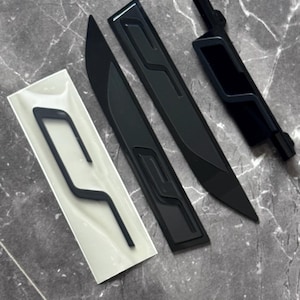 Könnte beinhalten: Eine Sammlung schwarzer Autoteile, darunter zwei lange, gebogene Teile mit geprägtem Design, ein kleineres Teil und ein Teil in einer transparenten Kunststoffverpackung. Die Teile sind auf einer grau marmorierten Oberfläche angeordnet.