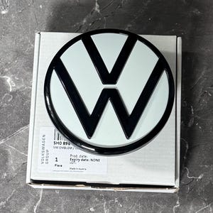 Könnte beinhalten: Ein kreisförmiges Volkswagen-Emblem mit einem schwarzen V- und W-Logo auf weißem Hintergrund, das auf einem weißen Karton liegt. Das Etikett auf der Schachtel zeigt den Text VOLKSWAGEN GROUP, 5H0 896, VW EMBLEM, 1 Piece, Prod. date, Expiry date: NONE, LOT, und Made In Austria.