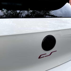 Puede incluir: Primer plano de la parte trasera de un coche blanco con un emblema negro y un logotipo "CS" magenta. La ventana trasera del coche es visible encima del emblema. La pintura del coche parece limpia.