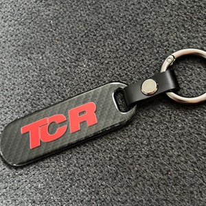 Peut inclure: Un porte-clés TCR noir et rouge avec un motif en fibre de carbone. Le porte-clés est doté d'une lanière en cuir noir et d'un anneau métallique argenté pour attacher les clés. Les lettres "TCR" sont en rouge.
