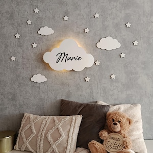 May include: White wooden cloud night light with the name "Marie" written in black script. The light is surrounded by small white wooden stars on a gray textured wall. A teddy bear with a wooden sign that says "Hallo Welt" is sitting on a bed with pillows.