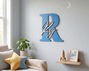 Op de afbeelding: Een grote blauwe houten letter R aan de muur, met de naam Rey in een lichtgekleurd cursief lettertype in het midden. De muur is versierd met witte stickers van een halve maan, sterren en sterrenbeelden rondom de letter.