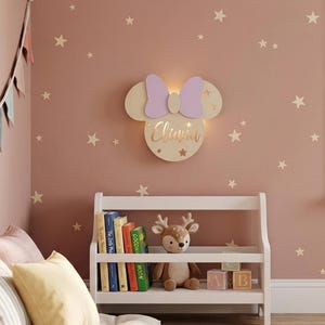 Può includere: Una lampada da parete a forma di testa di topo con un fiocco viola chiaro e il nome Eliana scritto in corsivo dorato. La luce emette un bagliore soffuso da dietro la struttura in legno, decorata con piccoli intagli a forma di stella.