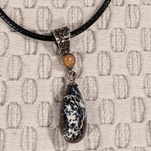 May include: A black cord necklace with a silver pendant featuring a teardrop-shaped opal with black and white patterns and a smaller opal bead above it.