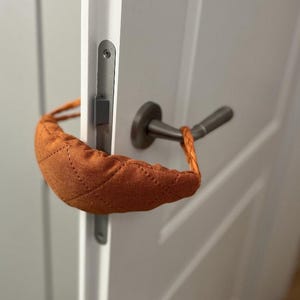 Può includere: Un fermaporta marrone con una copertura in tessuto trapuntato e un manico in corda. Il fermaporta è attaccato a una porta bianca con una maniglia della porta argentata.