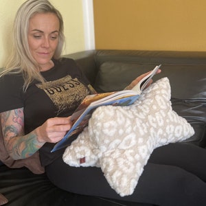 May include: A woman is sitting on a sofa reading a book while holding a white and beige star-shaped cushion with a leopard print.