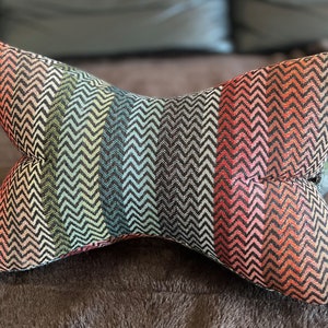 Op de afbeelding: Een kleurrijk kussen met een chevron-patroon in tinten roze, oranje, groen, blauw en zwart. Het kussen is gevormd als een vlinderdas.