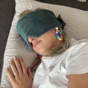May include: A teal sleep mask rests on a person's face, covering the eyes. The mask is made of a soft fabric. The person is wearing colorful beaded earrings. The person is wearing a white t-shirt.