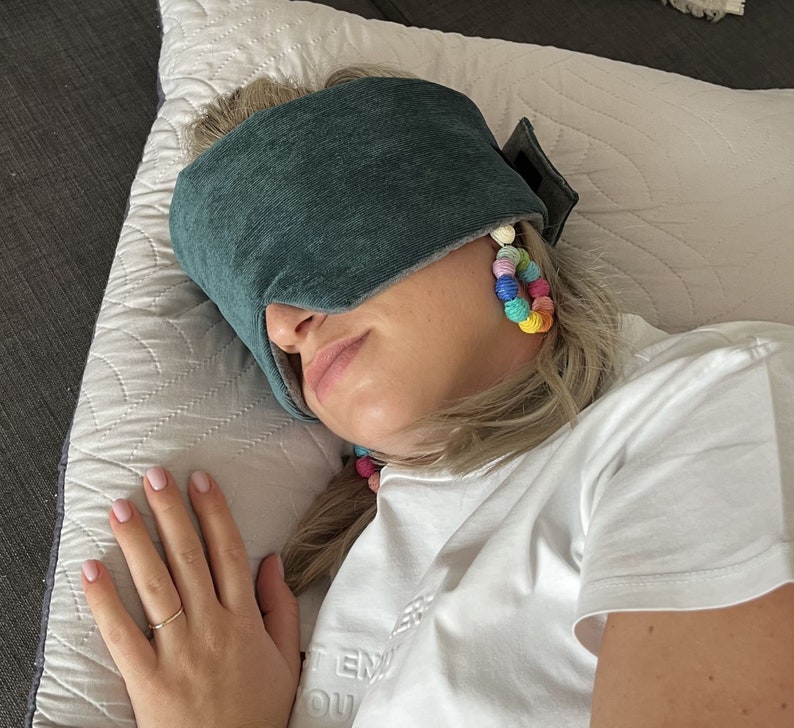 May include: A teal sleep mask rests on a person's face, covering the eyes. The mask is made of a soft fabric. The person is wearing colorful beaded earrings. The person is wearing a white t-shirt.