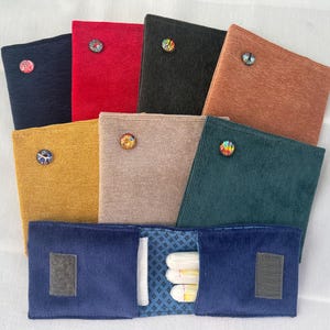 Könnte beinhalten: Eine Sammlung farbenfroher Cord-Stoffbeutel, jeweils mit einem dekorativen Knopf. Die Beutel sind in verschiedenen Farben erhältlich, darunter Marineblau, Rot, Schwarz, Senfgelb, Beige, Rost und Türkis. Ein Beutel ist geöffnet und zeigt Tampons.