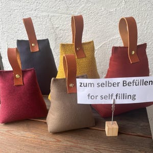Könnte beinhalten: Eine Auswahl an Stoff-Türstoppern in verschiedenen Farben, darunter Rot, Marineblau, Gelb und Braun. Jeder Türstopper hat einen Ledergriff, der mit einer Messingniete befestigt ist. Das Schild lautet "zum selber Befüllen for self filling."