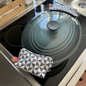 Op de afbeelding: Een teal Le Creuset braadpan met een zwarte knop staat op een kookplaat. Twee grijze en witte geometrisch patroon pannenlappen liggen op de pot. Eén pannenlap heeft een roze en gouden label met "Handmade".