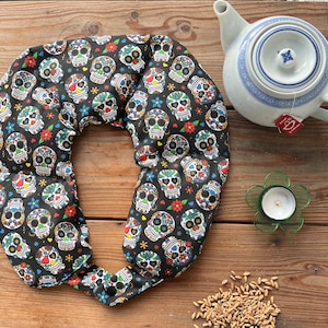 Könnte beinhalten: Ein schwarzer Nackenwärmer mit einem bunten Zuckerschädelmuster. Daneben eine weiße Teekanne mit blauem Design, ein grüner blütenförmiger Kerzenhalter und Getreide auf einer Holzoberfläche.