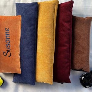 Könnte beinhalten: Eine Auswahl an Cordstoffartikeln in verschiedenen Farben: Orange mit dem Namen "Susanne" bestickt, Marineblau, Gelb, Burgunderrot und Braun. Zwei Spielzeugautos, eines gelb und eines schwarz, liegen daneben.