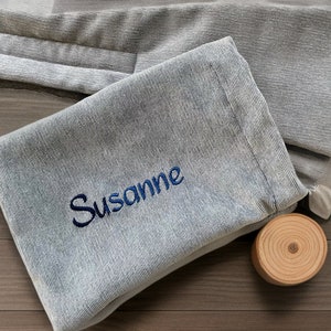 May include: A light blue, textured fabric pouch with the name "Susanne" embroidered in dark blue. A small, round wooden coaster sits beside the pouch. The pouch appears to be folded and has a soft, casual appearance.