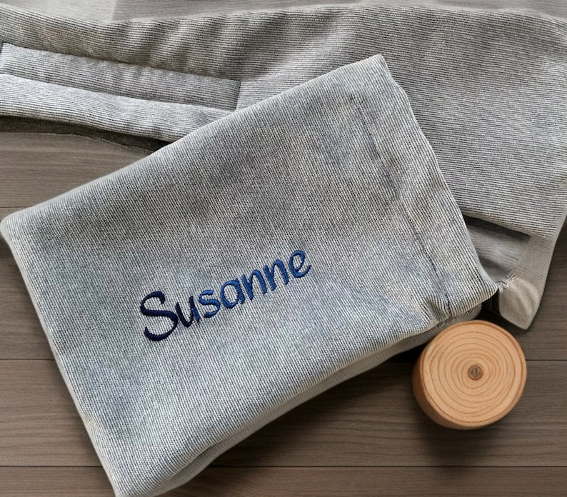 May include: A light blue, textured fabric pouch with the name "Susanne" embroidered in dark blue. A small, round wooden coaster sits beside the pouch. The pouch appears to be folded and has a soft, casual appearance.
