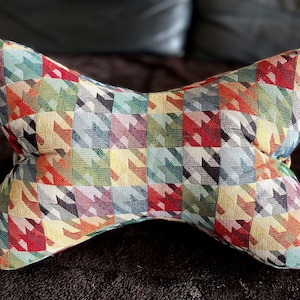 Op de afbeelding: Een kleurrijk, gepatroneerd kussen in de vorm van een vlinderdas. Het kussen is gemaakt van geweven stof met een geometrisch ontwerp in tinten rood, groen, geel, blauw en zwart.