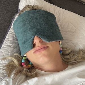 May include: A person wearing a teal eye mask, resting on a white pillow. The eye mask is made of a soft, textured fabric. The person is also wearing colorful beaded earrings. The background includes a gray couch.