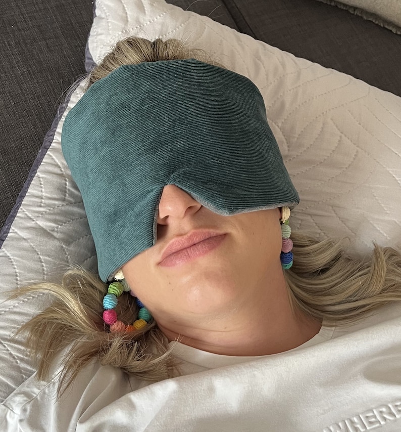 May include: A person wearing a teal eye mask, resting on a white pillow. The eye mask is made of a soft, textured fabric. The person is also wearing colorful beaded earrings. The background includes a gray couch.